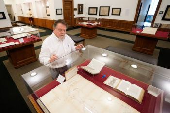 the curator stands next to a glass case containing a large map and several small books