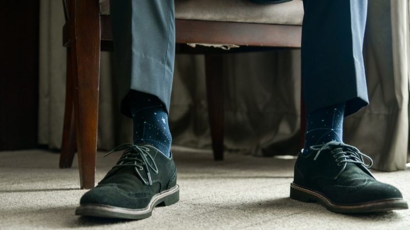 a close up photo of a man's feet and shins - he is sitting in a chair wearing black dress shoes and dark blue socks