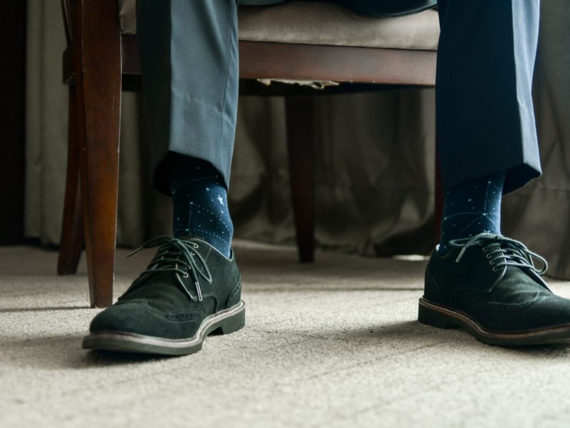a close up photo of a man's feet and shins - he is sitting in a chair wearing black dress shoes and dark blue socks