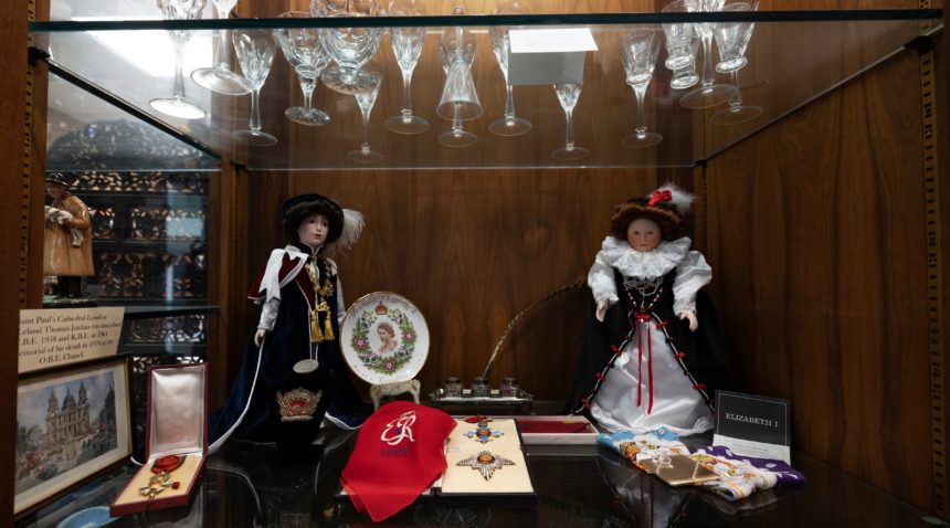 a photo of the interior of a glass display case showing badges, scarves, dolls, an inkwell, a commemorative plate, and other miscellaneous items
