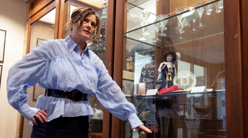 a photo of a woman in a blue shirt gestures towards a glass case containing various pins, medals, glassware and other items