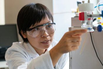 a photo of a woman in a lab coat and safety glasses looking at lab equipment