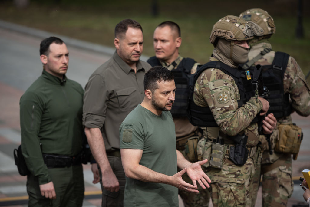 a photo of Ukrainian President Volodymyr Zelensky in front of a group of men, two of them in full military gear carrying rifles