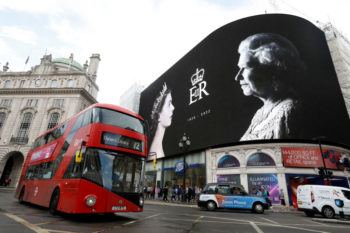 A tribute screen on a busy street in London shows black and white photos of the queen as a red bus drives by