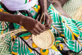 a close-up photo of the hands of a woman in a green and yellow patterned skirt sitting down and weaving a basket