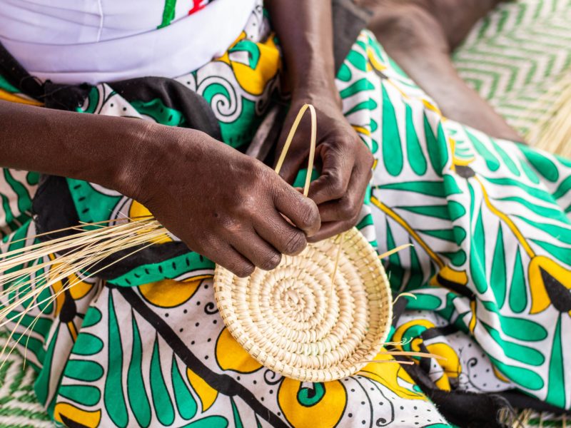 a close-up photo of the hands of a woman in a green and yellow patterned skirt sitting down and weaving a basket
