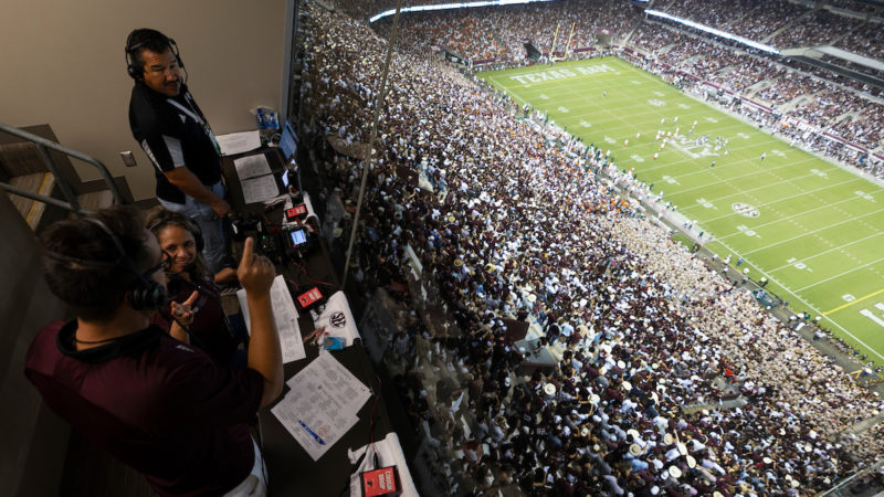 in a booth overlooking the crowd and football field at kyle field, two men and a woman stand in a broadcasting booth