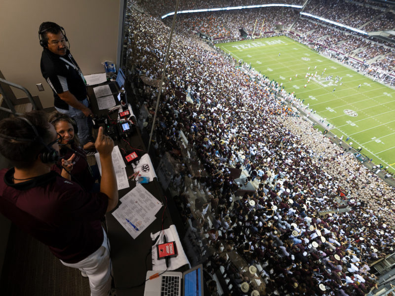 in a booth overlooking the crowd and football field at kyle field, two men and a woman stand in a broadcasting booth