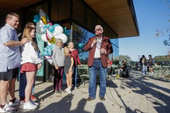 a man wearing a maroon jacket speaks into a microphone outside the creamery as family members look on