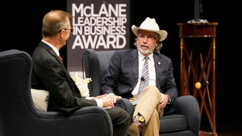 Beaver Aplin seated on a stage wearing a cowboy hat. A banner in the background reads 