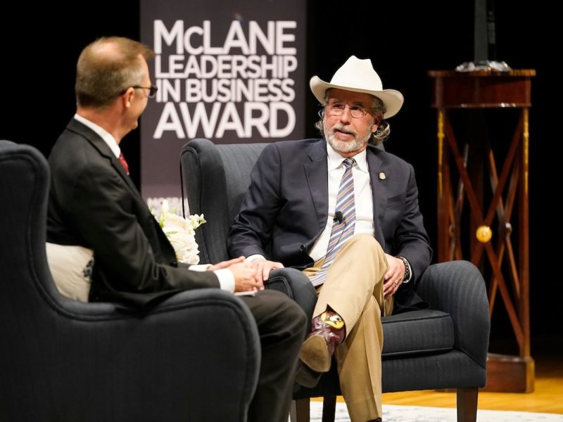 Beaver Aplin seated on a stage wearing a cowboy hat. A banner in the background reads 