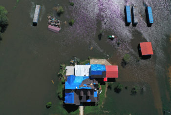 a top-down photo of a house sitting just above surrounding flood waters. The roof of the house has been patched up with blue tarps. Behind the house, a tractor, outbuilding and several storage containers sit in the floodwater.