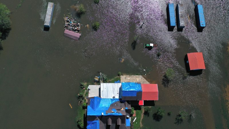 a top-down photo of a house sitting just above surrounding flood waters. The roof of the house has been patched up with blue tarps. Behind the house, a tractor, outbuilding and several storage containers sit in the floodwater.