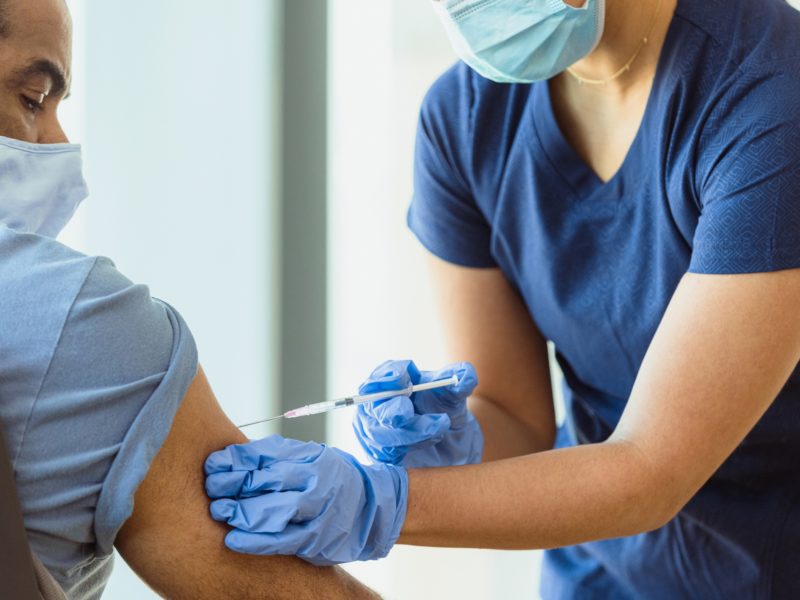 An unrecognizable adult female nurse gives her mature adult male patient a flu shot in his arm. He watches intently. Both the nurse and the patient wear protective masks.