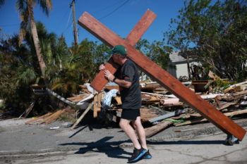 A man carries a large wooden cross over his shoulder through an area destroyed by a hurricane
