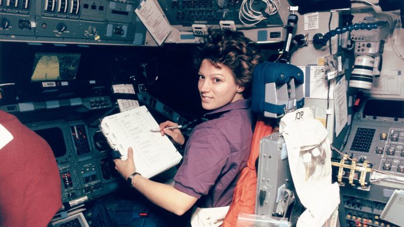 archival photo of astronaut eileen collins sitting down inside a space shuttle