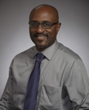 a portrait photo of a man with glasses and a short mustache/goatee in a grey shirt and purple tie