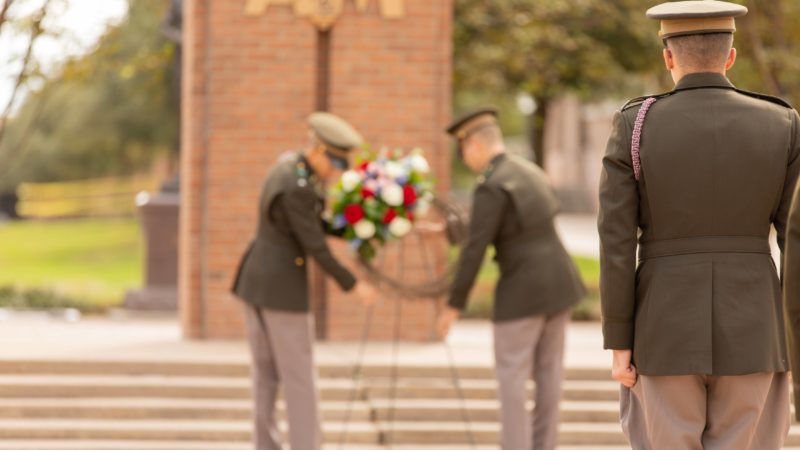 A cadet watches on as two cadets in the background set up a wreath in front of the Corps arches on campus