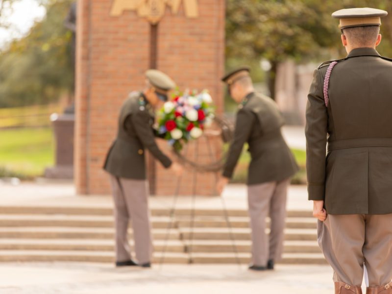A cadet watches on as two cadets in the background set up a wreath in front of the Corps arches on campus