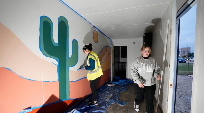 foto dua wanita muda melepas selotip dari mural yang dicat di dalam klinik kontainer pengiriman