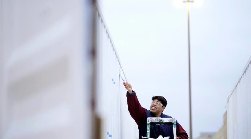 a photo of a young man in a dark blue and maroon jacket standing on a ladder applying white paint to the side of a shipping container