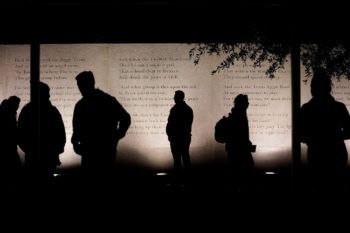 People in silhouette standing in front of a wall engraved with the poem "The Last Corps Trip."