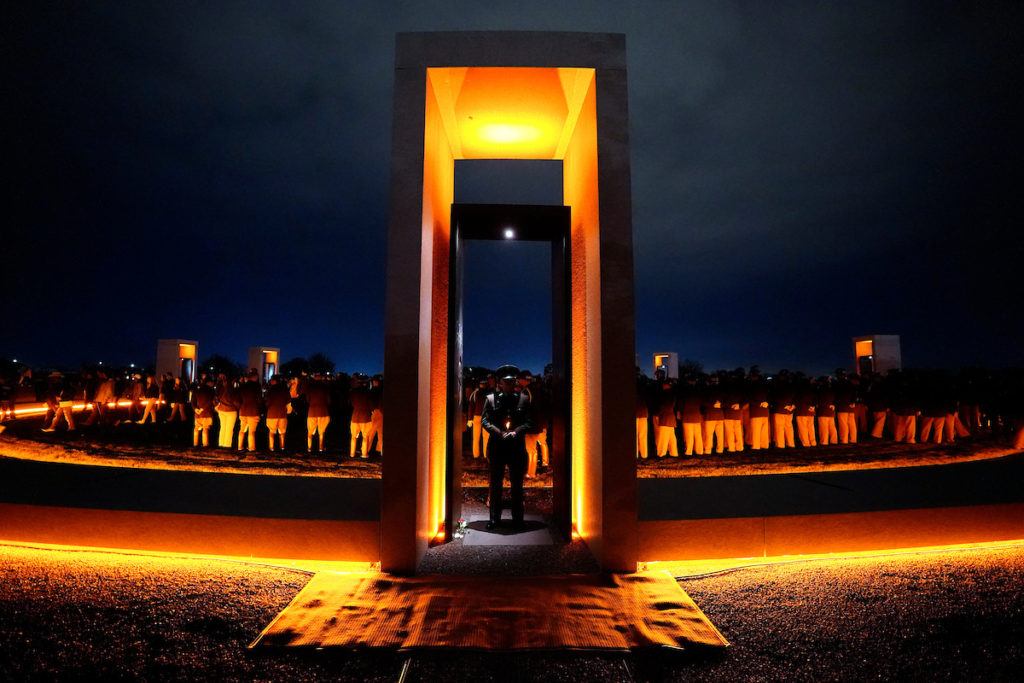 A photo of the bonfire memorial illuminated at night with a cadet standing in a portal. In the background, other cadets are standing at attention.