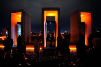 Three rectangular portals are illuminated in golden light. A cadet stands in each, while people surround the memorial in the darkness.