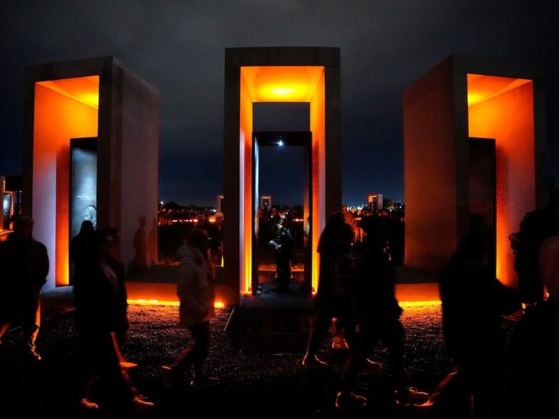 Three rectangular portals are illuminated in golden light. A cadet stands in each, while people surround the memorial in the darkness.