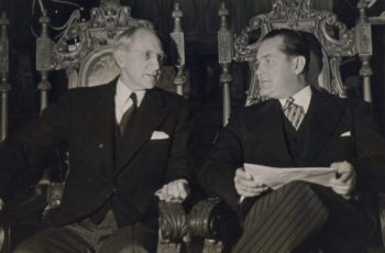 a photo of two men in suits sitting in ornately carved wooden chairs and carrying on a conversation