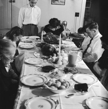 Black and white photo of a family sitting around a table praying. The table is set for Thanksgiving with a turkey in the center.