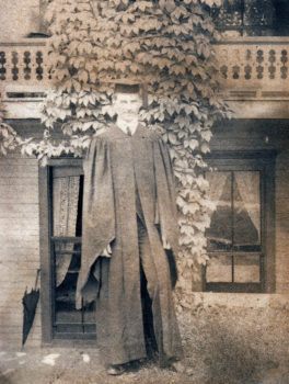 a photo of a young man with a slight smile in a graduation cap and gown. a cluster of ivy grows on the building behind him.