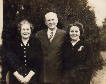 a photo of a man in a gray suit between an older and younger woman, both wearing black dresses. all are smiling.