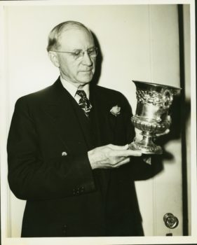 a photo of an older man in glasses holding a large metal trophy cup