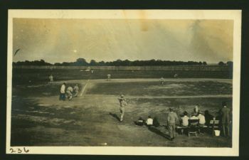 a photo of a game of baseball being played on a sparse field with a simple fence and lots of trees in the background