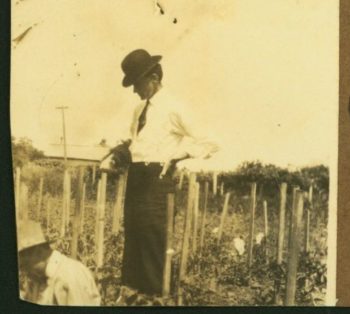 a photo of a man in a white shirt, black tie, and black bowler hat standing in a field with his hands on his hips looking at some crops