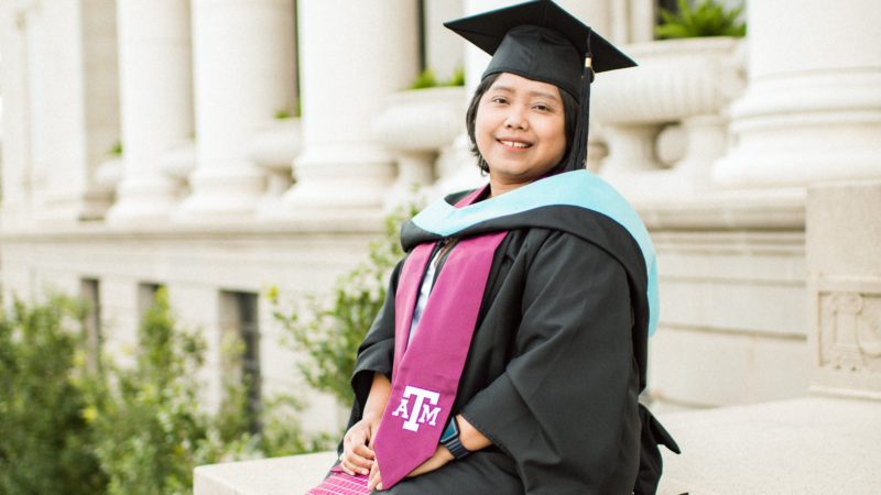 a photo of a woman in a dress, heels, and a graduation cap and gown sitting outside the administration building on the Texas A&M campus