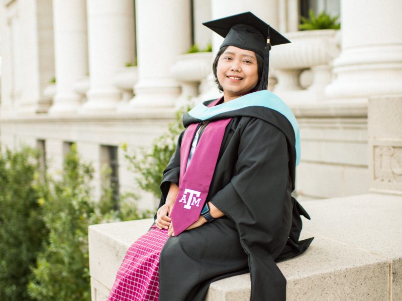a photo of a woman in a dress, heels, and a graduation cap and gown sitting outside the administration building on the Texas A&M campus