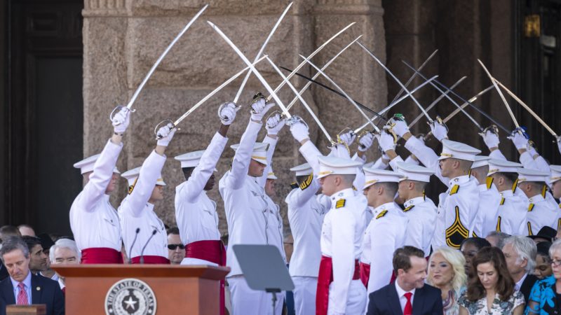 a photo of cadets in white uniforms holding up sabers to form an archway, creating a path to a podium in front of the state capitol building