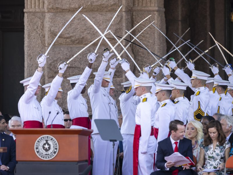 a photo of cadets in white uniforms holding up sabers to form an archway, creating a path to a podium in front of the state capitol building