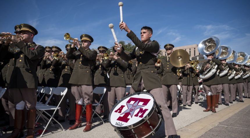 a photo of uniformed cadets playing instruments at the state capitol. in the center of the photo is a bass drummer with his hat off and his arms raised