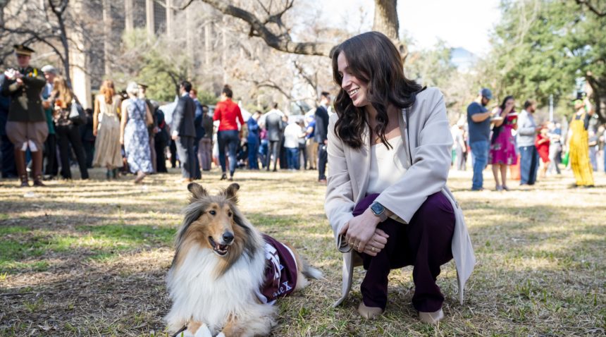 a photo of reveille laying on the grass in the shade next to a smiling woman in business casual attire