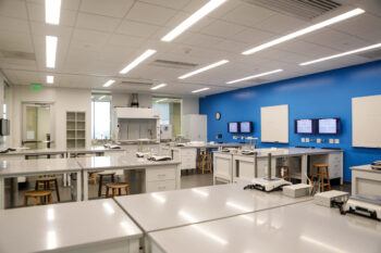 a photo of a chemistry lab room with a blue wall decked out with white boards and computer monitors. rows of lab tables and stools fill the majority of the room, with a large fume hood in the back