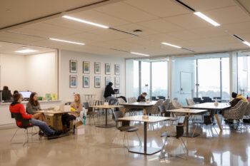 a photo of students sitting at tables in front of glass study rooms and a large service desk area recessed into the wall behind them