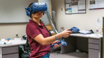 una foto di una donna con una polo marrone che utilizza un visore per realtà virtuale e controller
