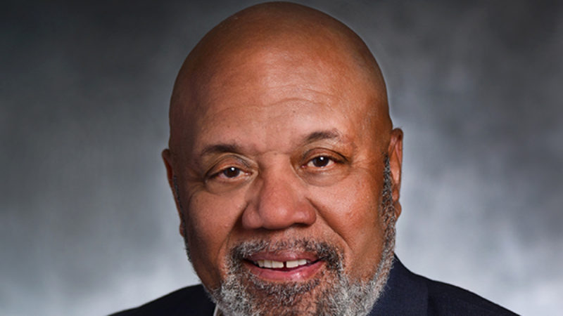 a photo of a man with a black and grey beard smiling into the camera. he is wearing a navy blue sports coat over a white button-up shirt.