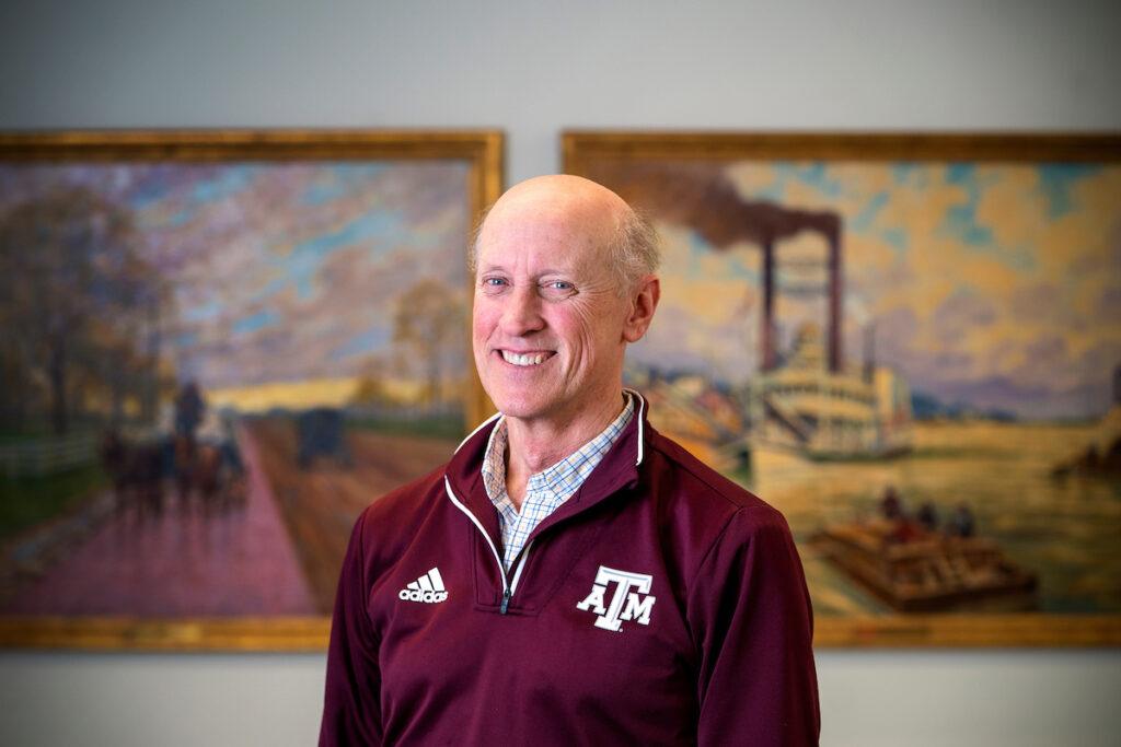 a photo of a man standing in front of two paintings and smiling. he is wearing a maroon Adidas Texas A&M quarter-zip pullover over a plaid button-up shirt.