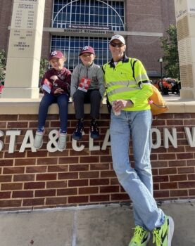 a photo of a man in a bright yellow safety shirt next to two small children in Texas A&M caps in front of Kyle Field