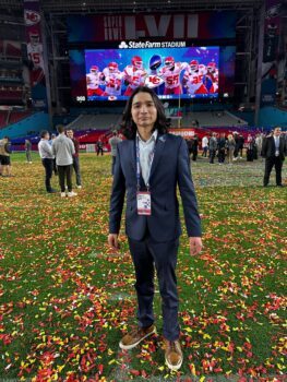 a photo of a man in a navy blue suit with long dark hair lookin into the camera as he stands on a football field covered in yellow red and white confetti.