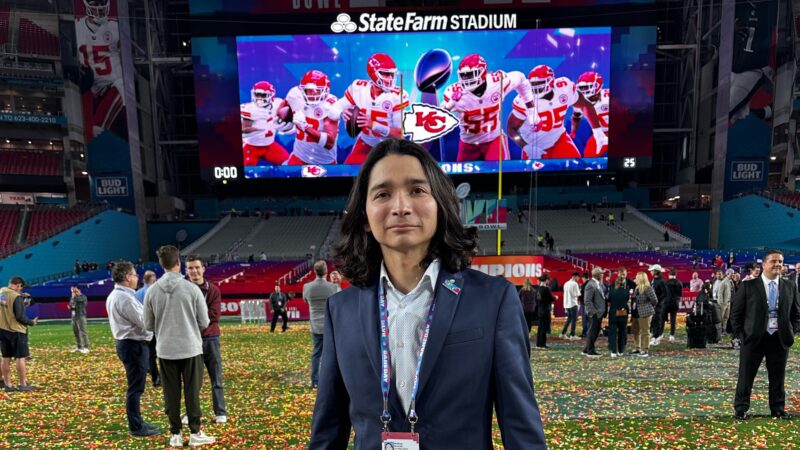 a photo of a man in a navy blue suit with long dark hair lookin into the camera as he stands on a football field covered in yellow red and white confetti.
